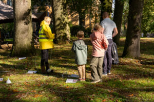 Kinderen en volwassenen buiten, omgeven door bomen en koorden op de grond, mogelijk een activiteit.