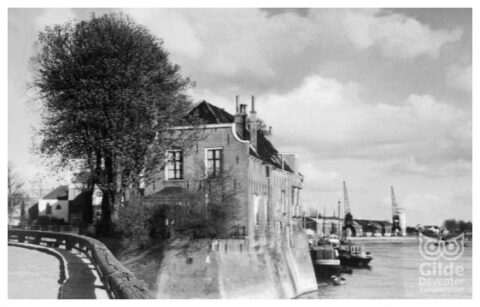 Oude foto van een muurhuis aan de waterkant, met bomen en schepen op de achtergrond.