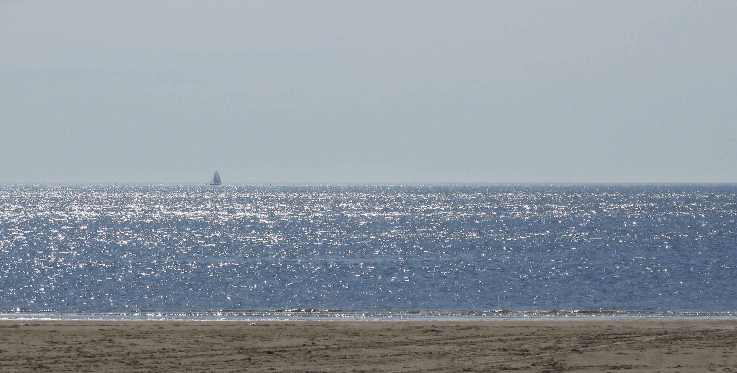 Zee met fonkelend water en een zeilboot aan de horizon, strand op de voorgrond.