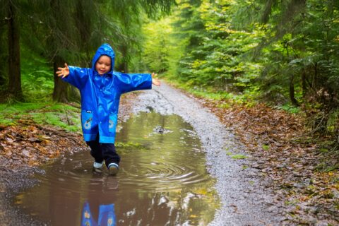 Een kind in blauwe regenjas speelt in een plas op een bospad.