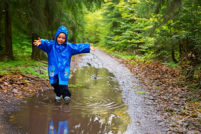 Een kind in blauwe regenjas speelt in een plas op een bospad.