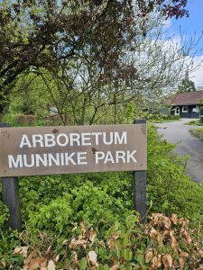 Bord van Arboretum Munnike Park omgeven door groene struiken en bomen; pad en huisje op de achtergrond.