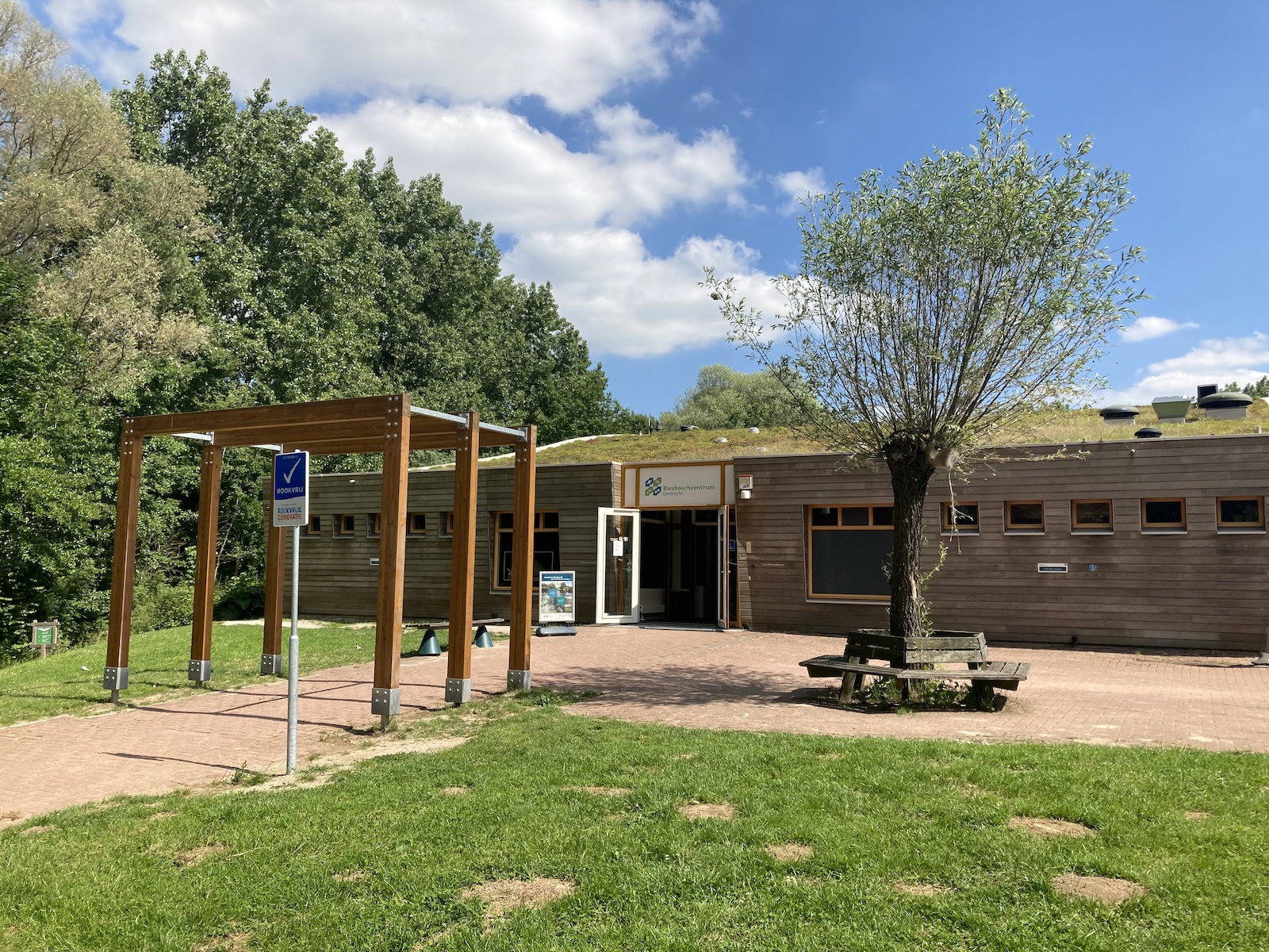 Houten gebouw met groendak, naast een parkeerbord en een zitbank rond een boom.