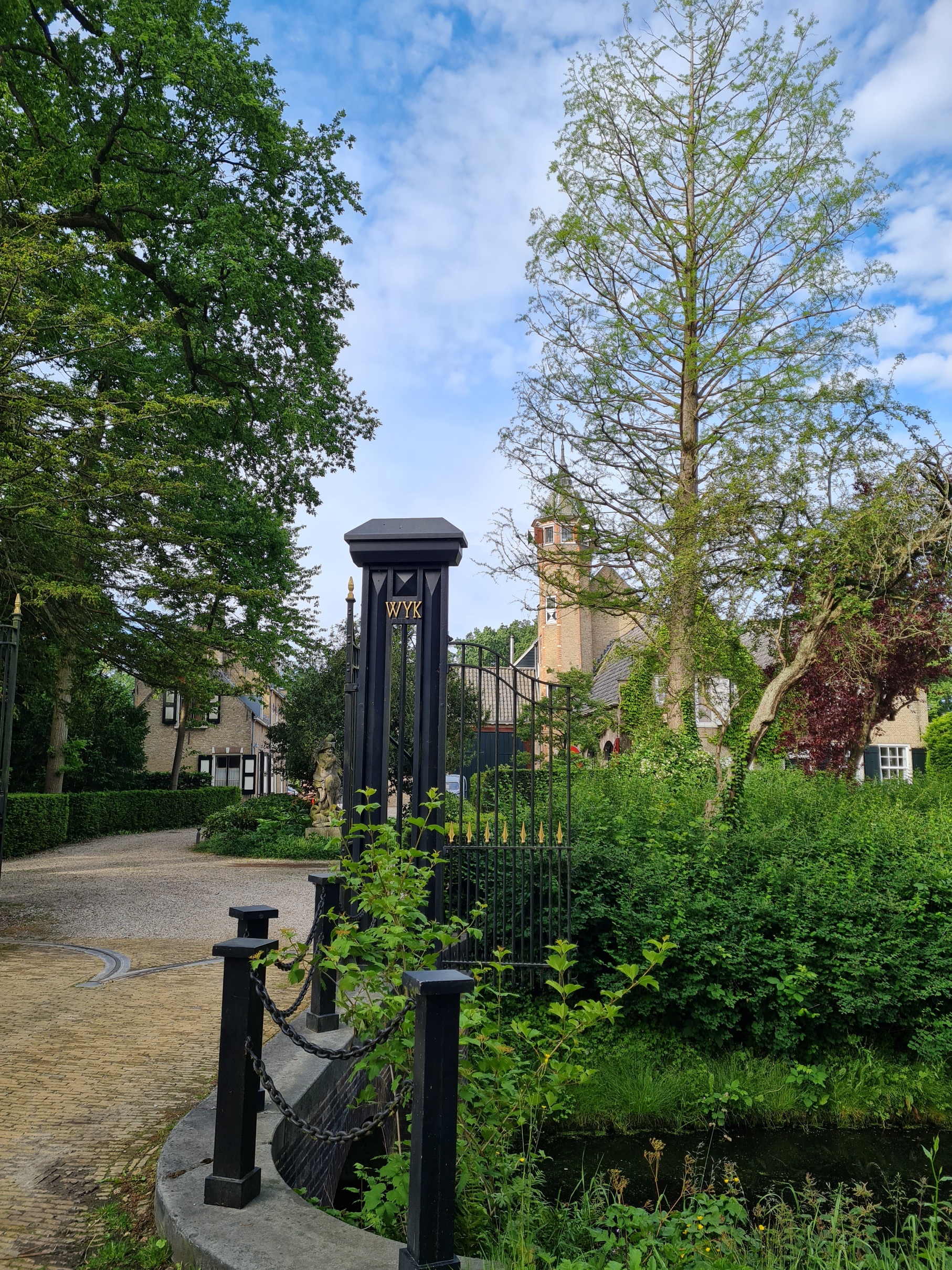 Smeedijzeren poort met groene bomen, een pad en een kerktoren op de achtergrond.