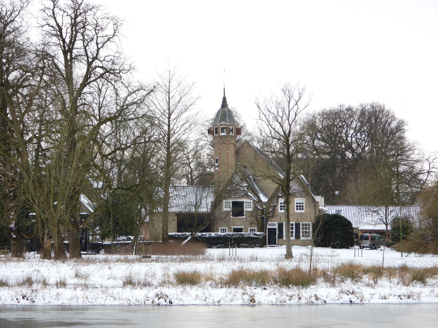 Sneeuw bedekt een historisch gebouw met toren te midden van kale bomen naast een bevroren rivier.