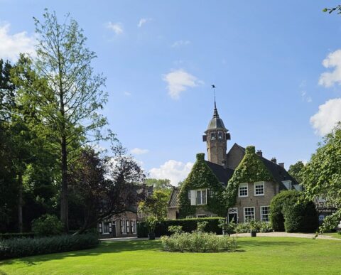 Historisch huis met torentje omgeven door groene tuin en vijver onder blauwe hemel.