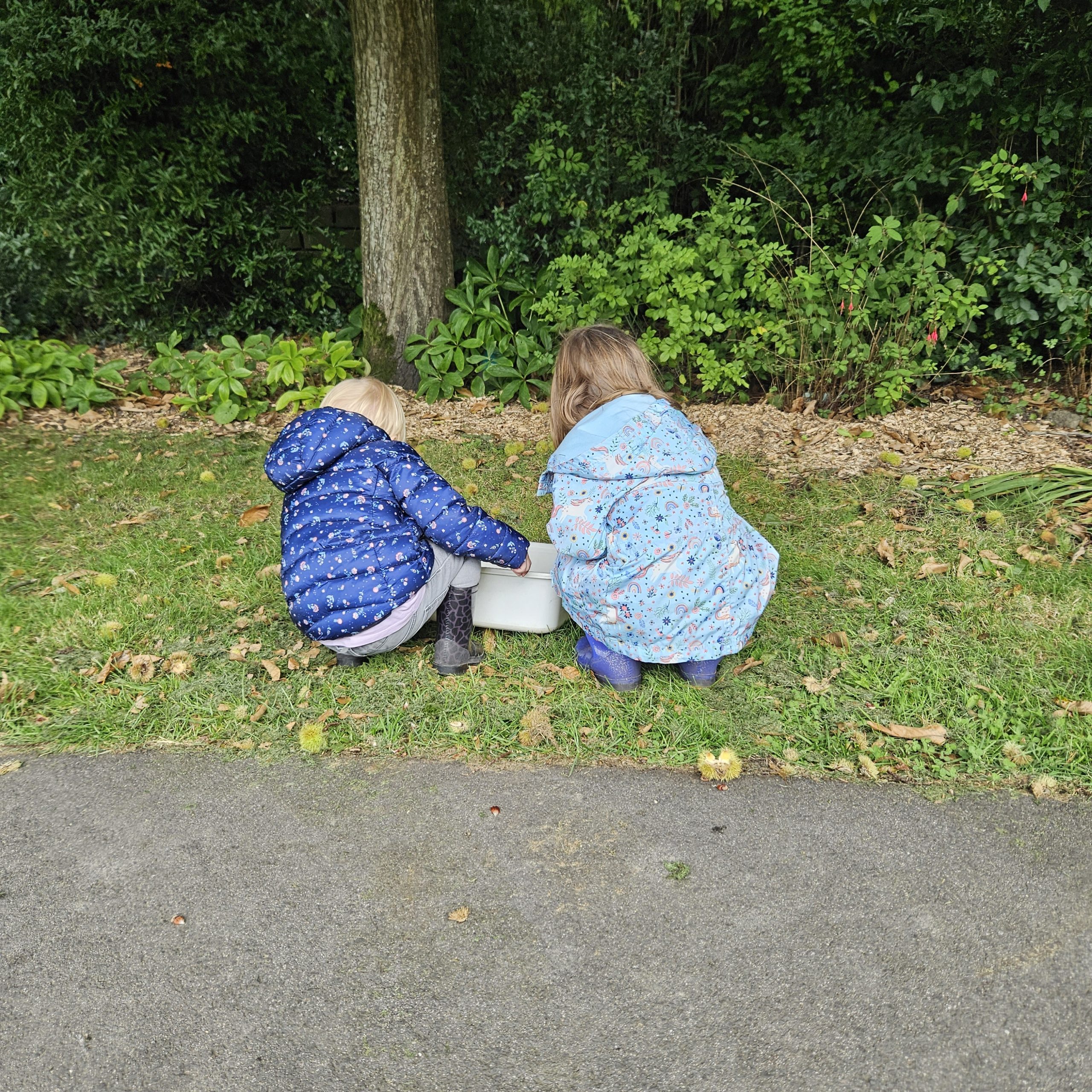 Twee kinderen in blauwe jassen verzamelen iets in een doos in een groene tuin.