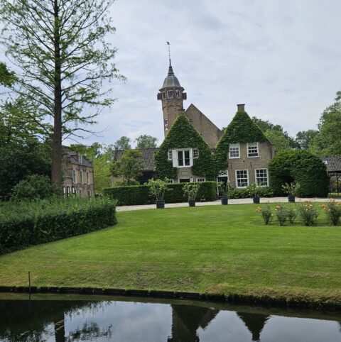 Huis met toren, omgeven door tuin en vijver, onder bewolkte lucht.