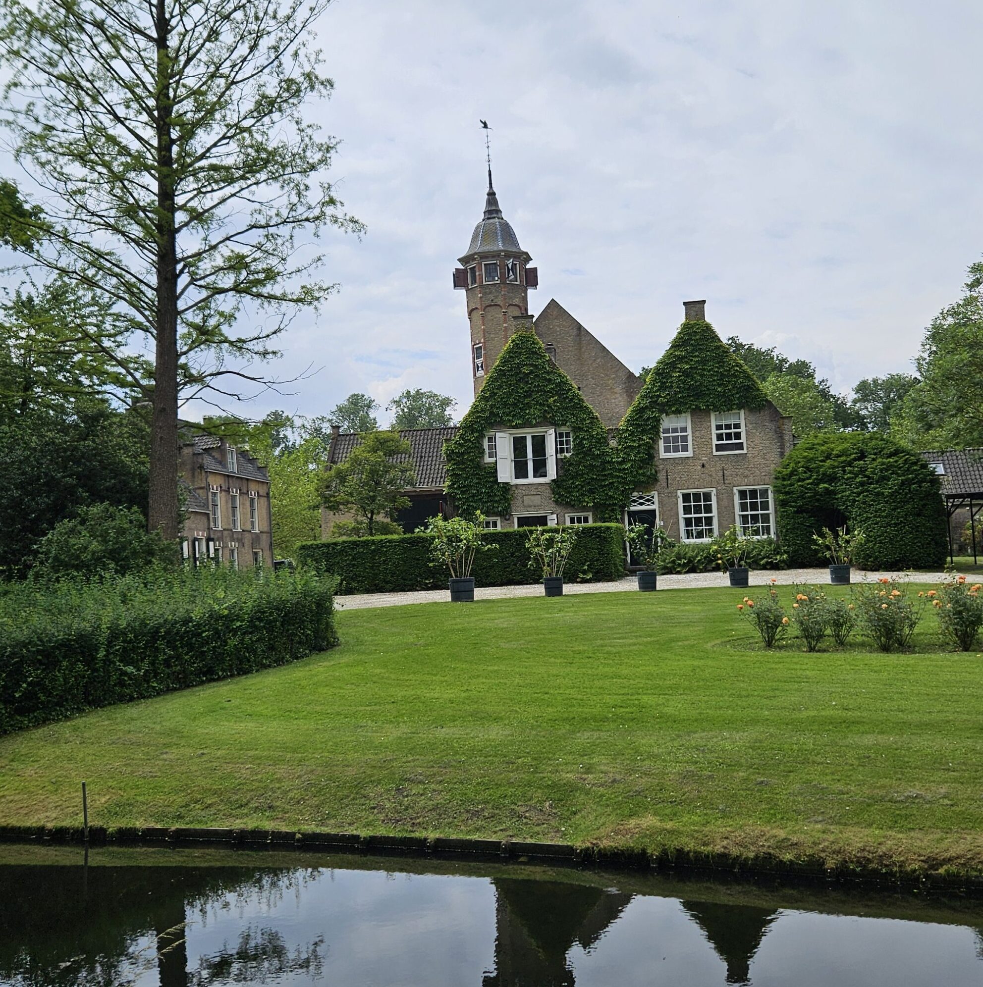 Huis met toren, omgeven door tuin en vijver, onder bewolkte lucht.