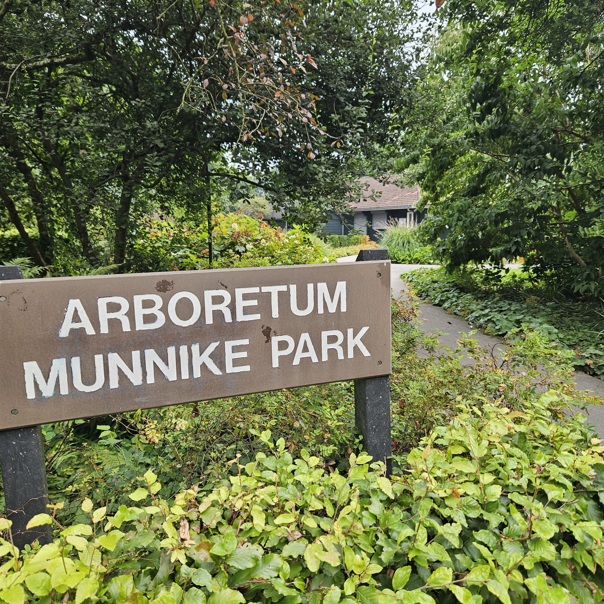 Bruin bord met tekst "Arboretum Munnike Park", omgeven door groene bomen en struiken.
