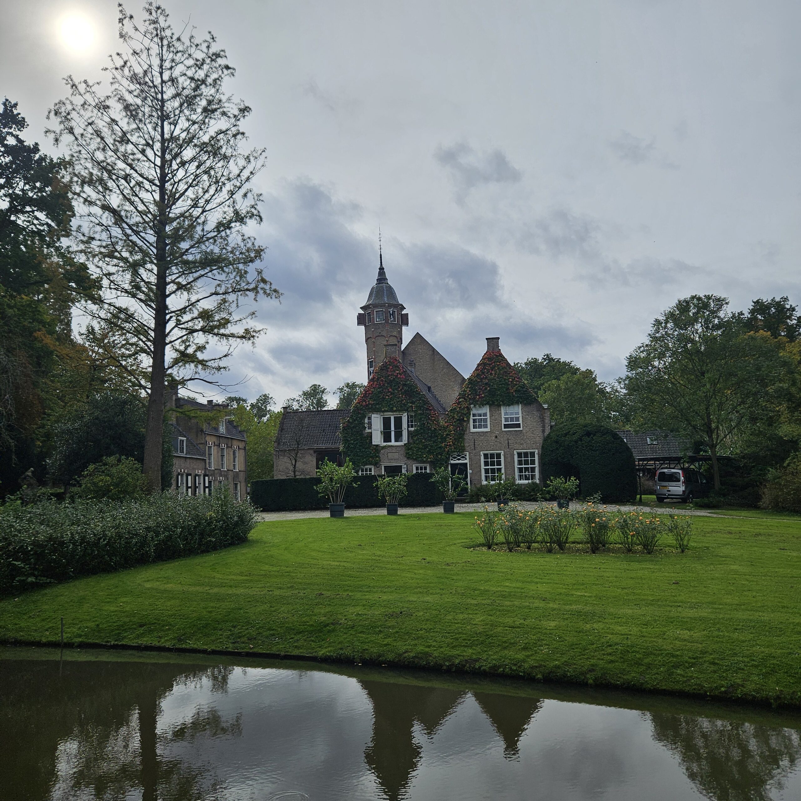 Historisch landhuis met toren, omgeven door groene tuin en vijver, onder bewolkte lucht.
