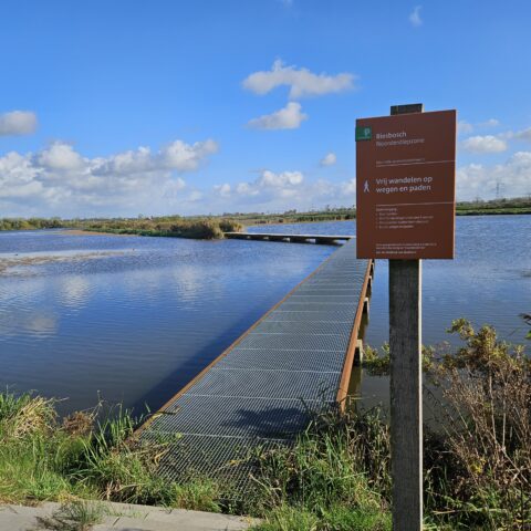 Brug over water met bord "Vrij wandelen", omringd door natuur en blauwe lucht.