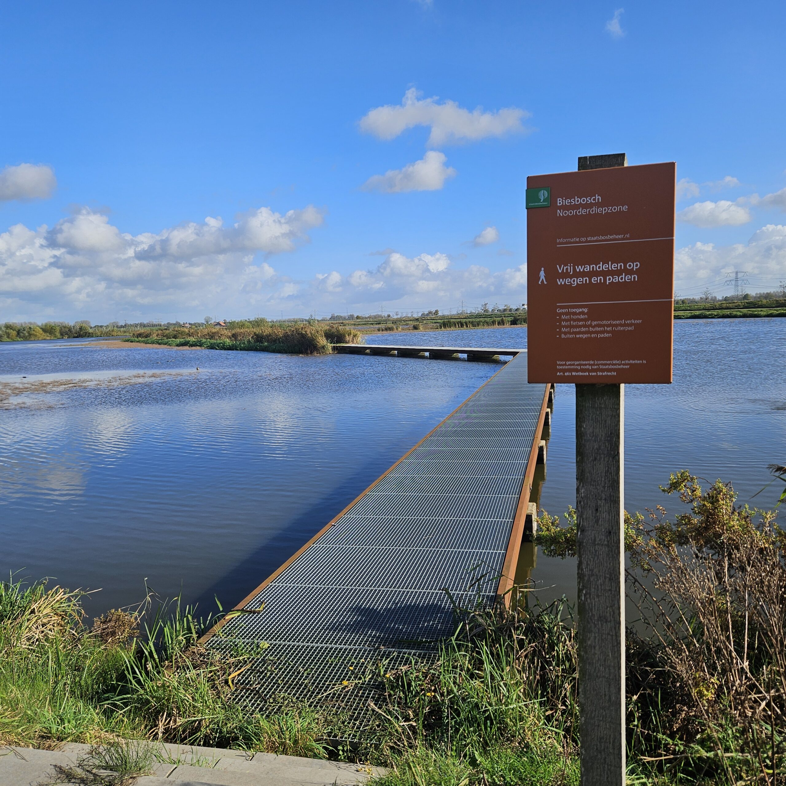Brug over water met bord "Vrij wandelen", omringd door natuur en blauwe lucht.