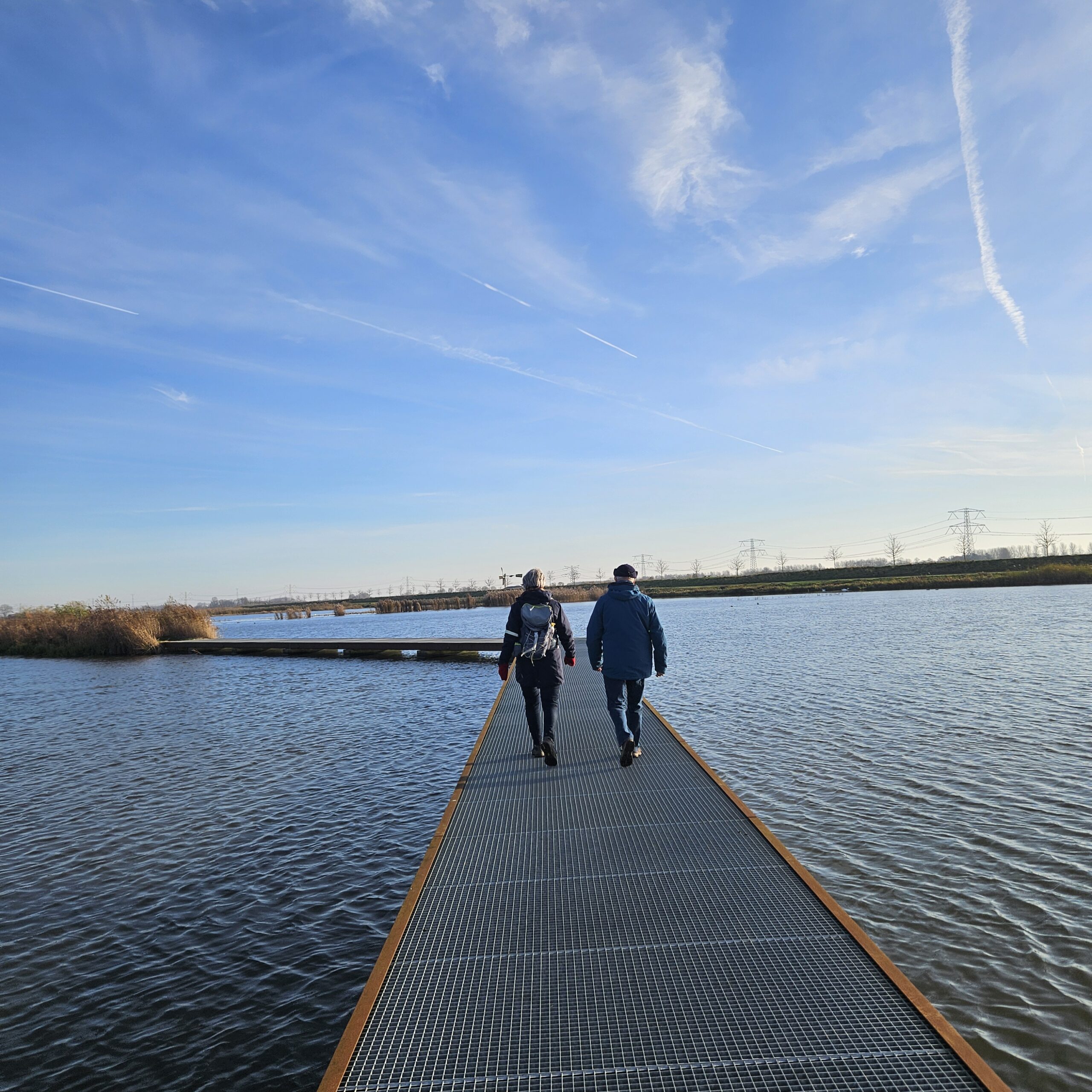 Twee personen wandelen op een metalen loopbrug over het water onder een heldere, blauw bewolkte hemel.
