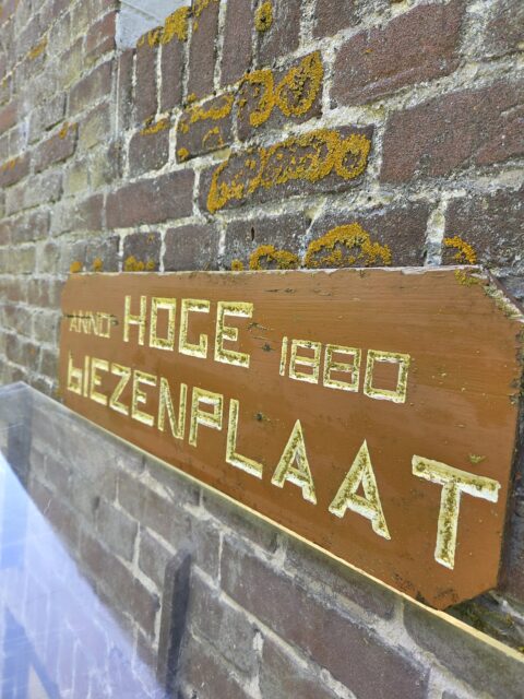 Bruine houten bord met gele tekst "Hoge Biezenplaat 1880" op een met mos begroeide bakstenen muur.
