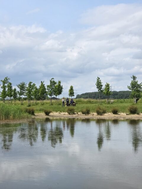 Drie mensen staan op een grasveld bij een vijver, omringd door bomen onder een bewolkte hemel.