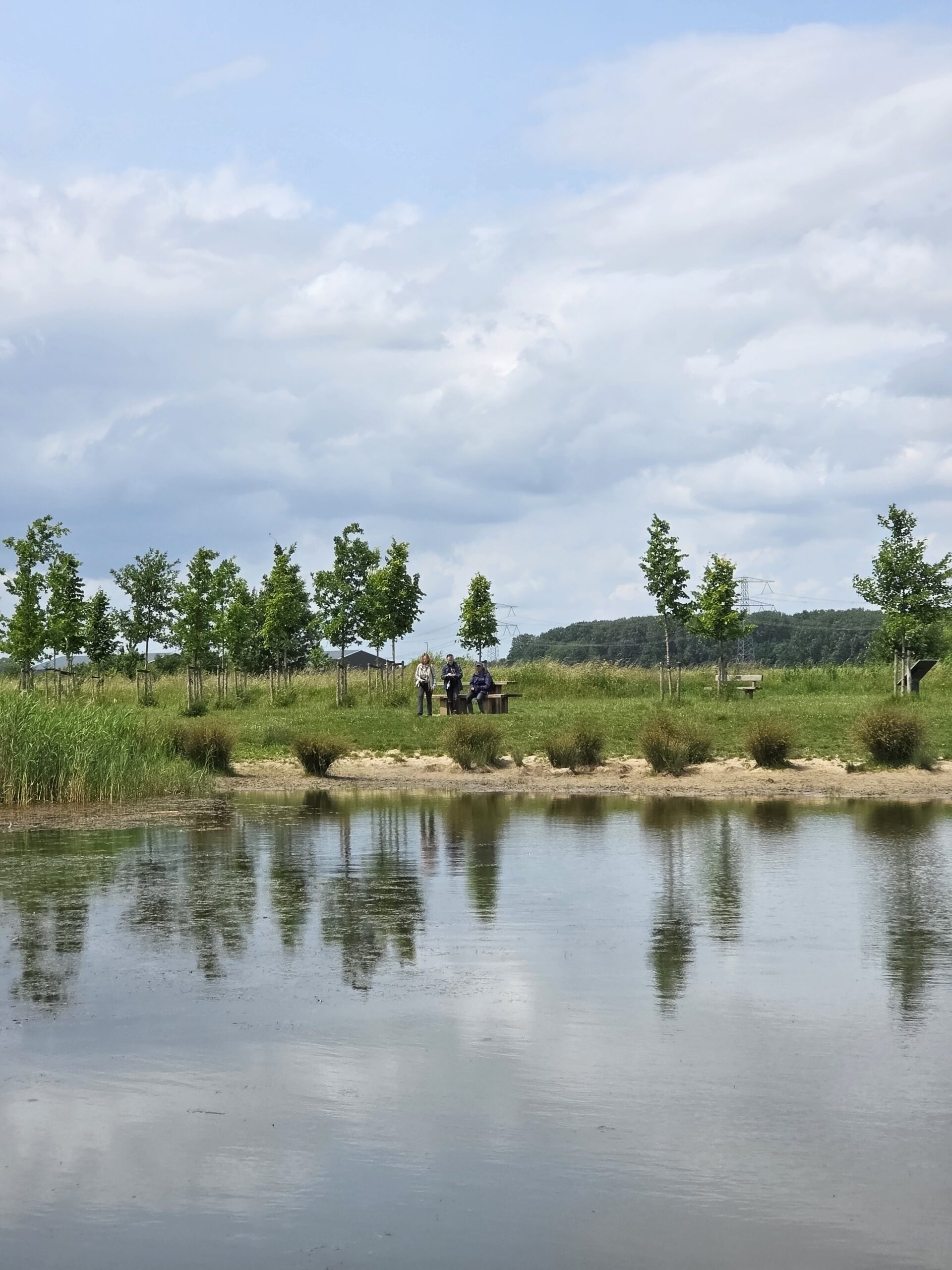 Drie mensen staan op een grasveld bij een vijver, omringd door bomen onder een bewolkte hemel.