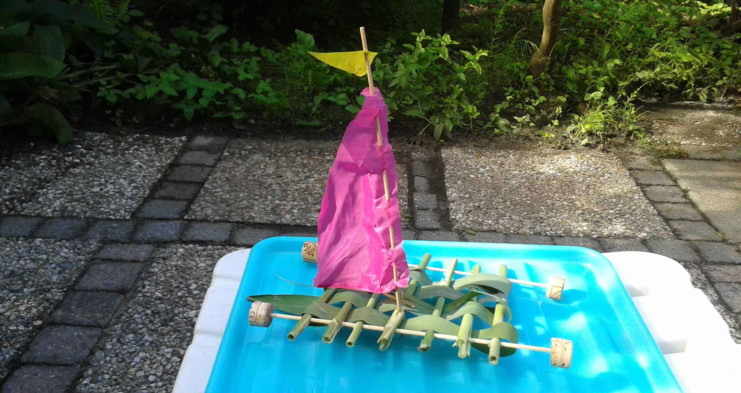 Klein vlot met roze zeil en kurken drijvers op een blauwe waterbak, buiten in een tuin.