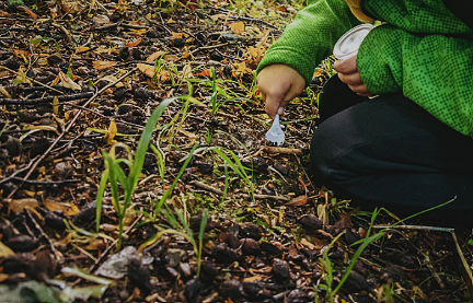 Kind graaft aarde om met plastic lepeltje, omgeven door gras en bladeren.