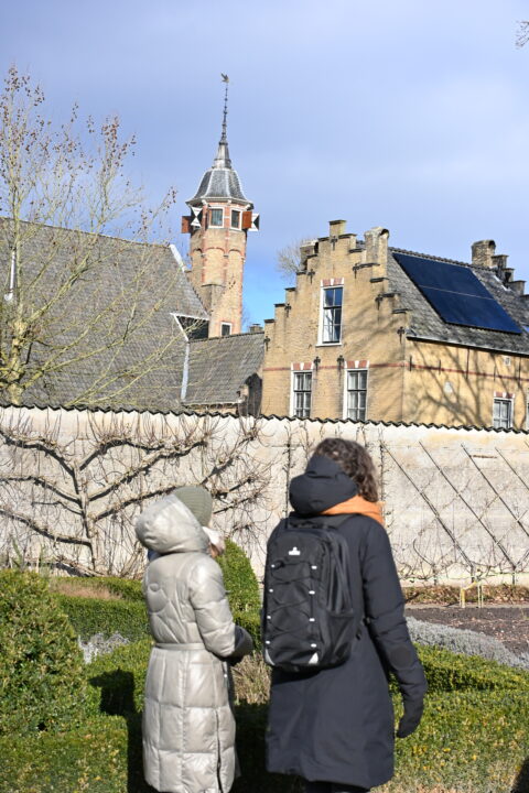 Twee mensen in winterkleding lopen bij een historisch gebouw met toren en zonnepanelen op het dak.