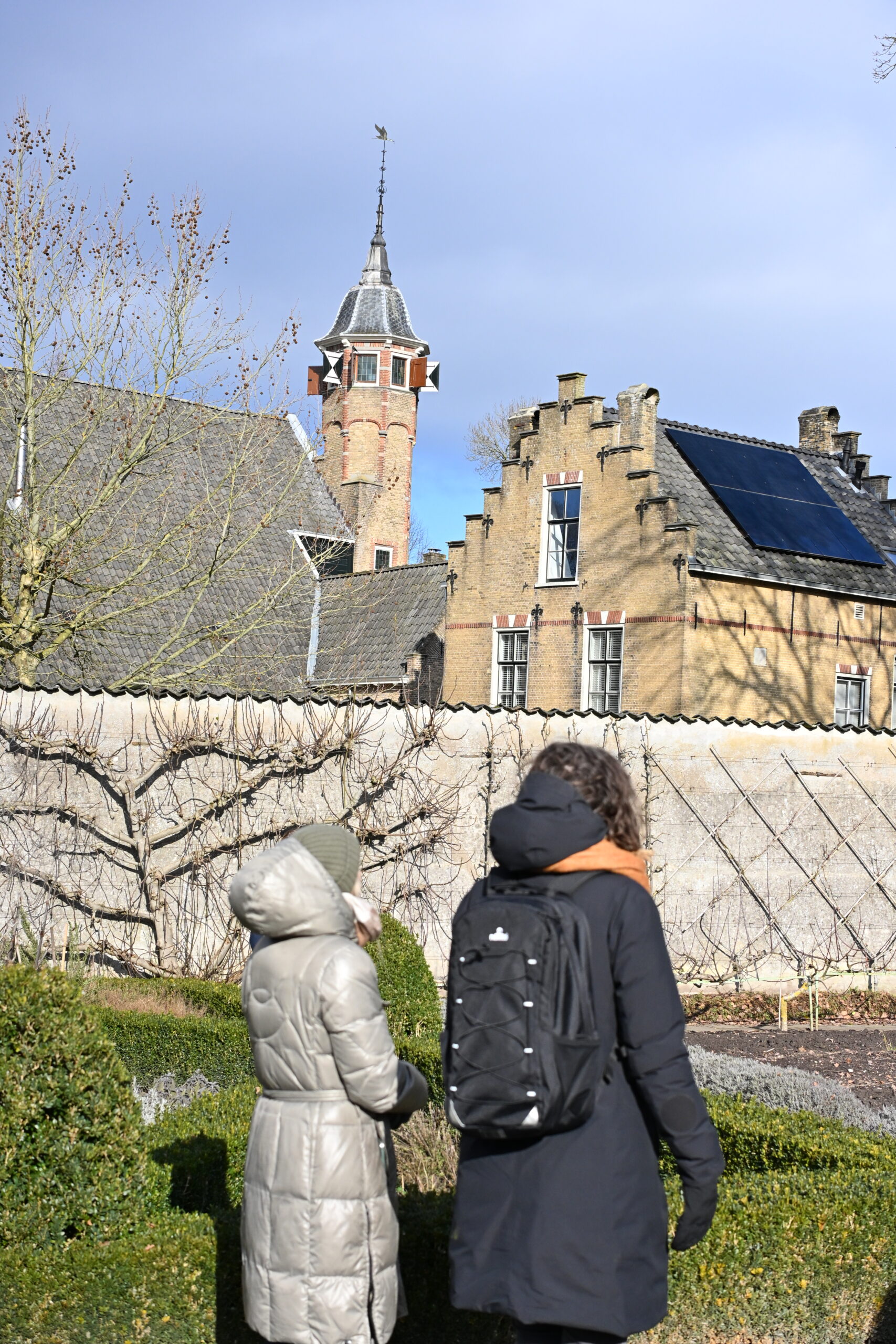 Twee mensen in winterkleding lopen bij een historisch gebouw met toren en zonnepanelen op het dak.