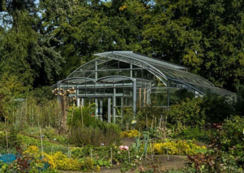 Glazen kas omringd door weelderige tuin met diverse planten en bomen op de achtergrond.