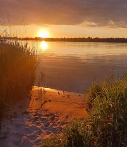 Zonsondergang boven een rustig meer, met zandstrand en gras in de voorgrond.