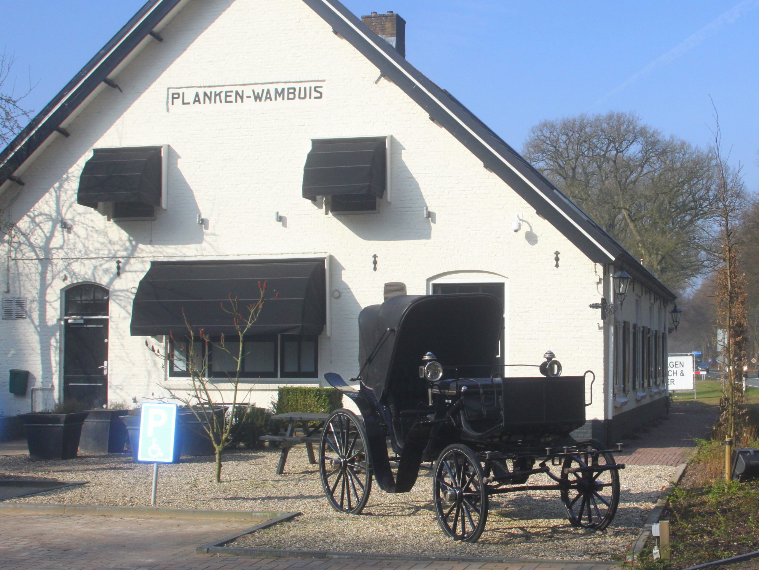 Wit gebouw "Planken Wambuis", oude koets vooraan, parkeerbord voor gehandicapten zichtbaar.