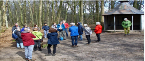 Groep mensen luistert naar een gids in een bos bij een kleine hut onder een zonnige hemel.