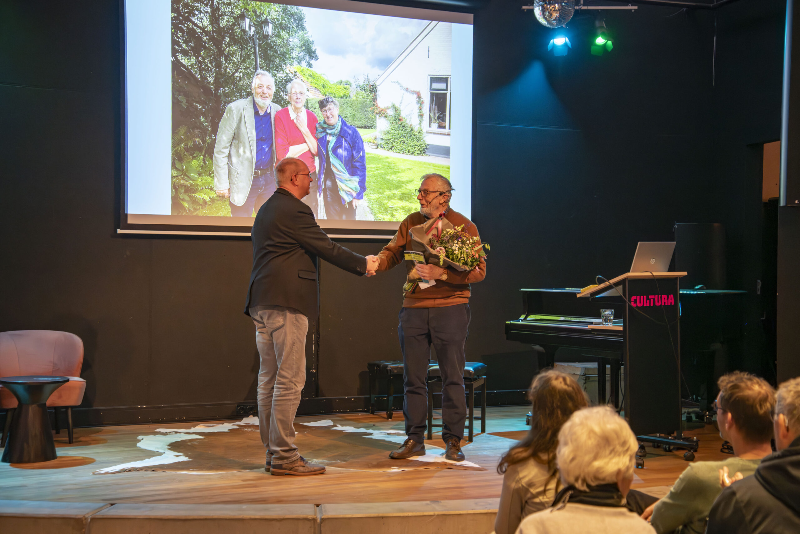 Twee mannen schudden handen op een podium, één met bloemen, publiek kijkt toe.