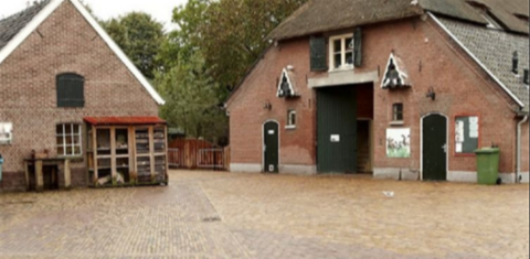 Historische boerenhof met twee bakstenen gebouwen, een binnenplaats en groene bomen op de achtergrond.