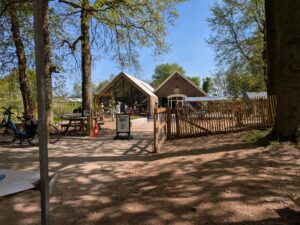 Buitenomgeving met een open café, fietsen, houten hek en bezoekers onder bomen.