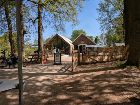 Buitenomgeving met een open café, fietsen, houten hek en bezoekers onder bomen.