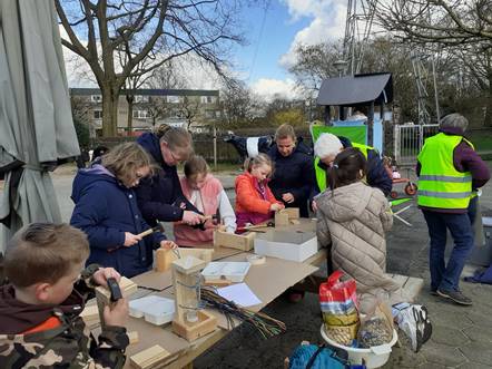 Kinderen en volwassenen knutselen samen aan een tafel buiten in een park onder een bewolkte hemel.