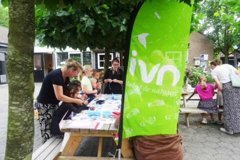 Kinderen knutselen aan een tafel buiten onder bomen, met een groene IVN-vlag.