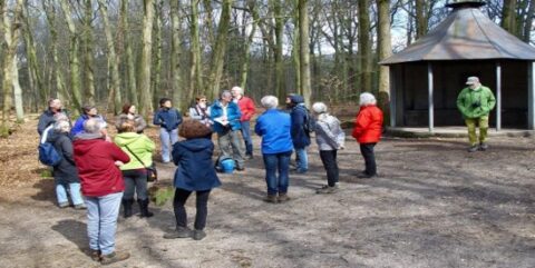 Een groep mensen staat buiten in een bos, in gesprek bij een houten hut.