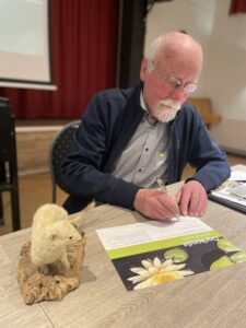 Oudere man schrijft op papier bij opgezette wezel, op tafel met infobrochure en rode gordijnen.
