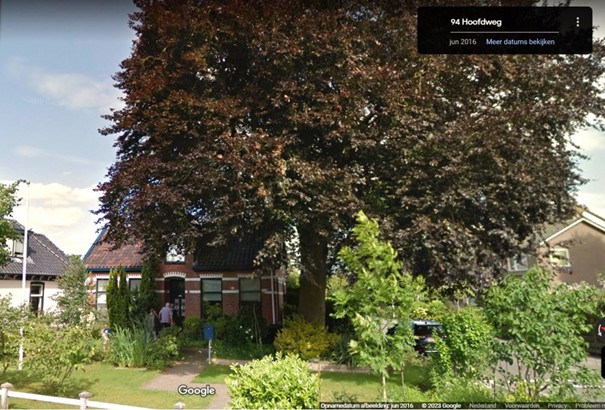 Voorgevel van een huis omringd door bomen en groen, met blauwe lucht op de achtergrond.