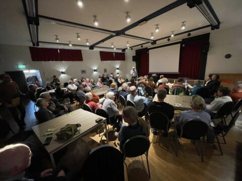 Een volle zaal met mensen die luisteren naar een spreker voor een groot scherm in een vergaderruimte.