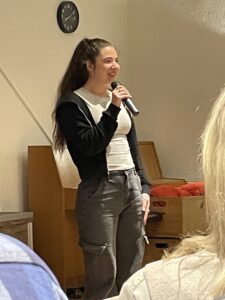 Vrouw met microfoon spreekt voor een groep; klok aan de muur.