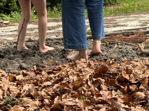 Twee mensen lopen blootsvoets over een modderige ondergrond, omringd door droge bladeren.