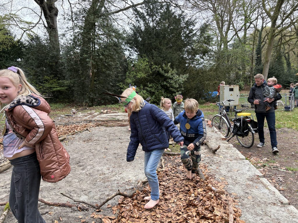 Kinderen lopen op blote voeten over bladeren in een bosrijke omgeving. Fietsen en volwassenen op achtergrond.