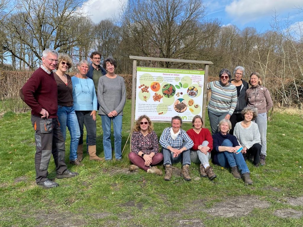 Een groep mensen poseert naast een informatiebord in een open veld met bomen.