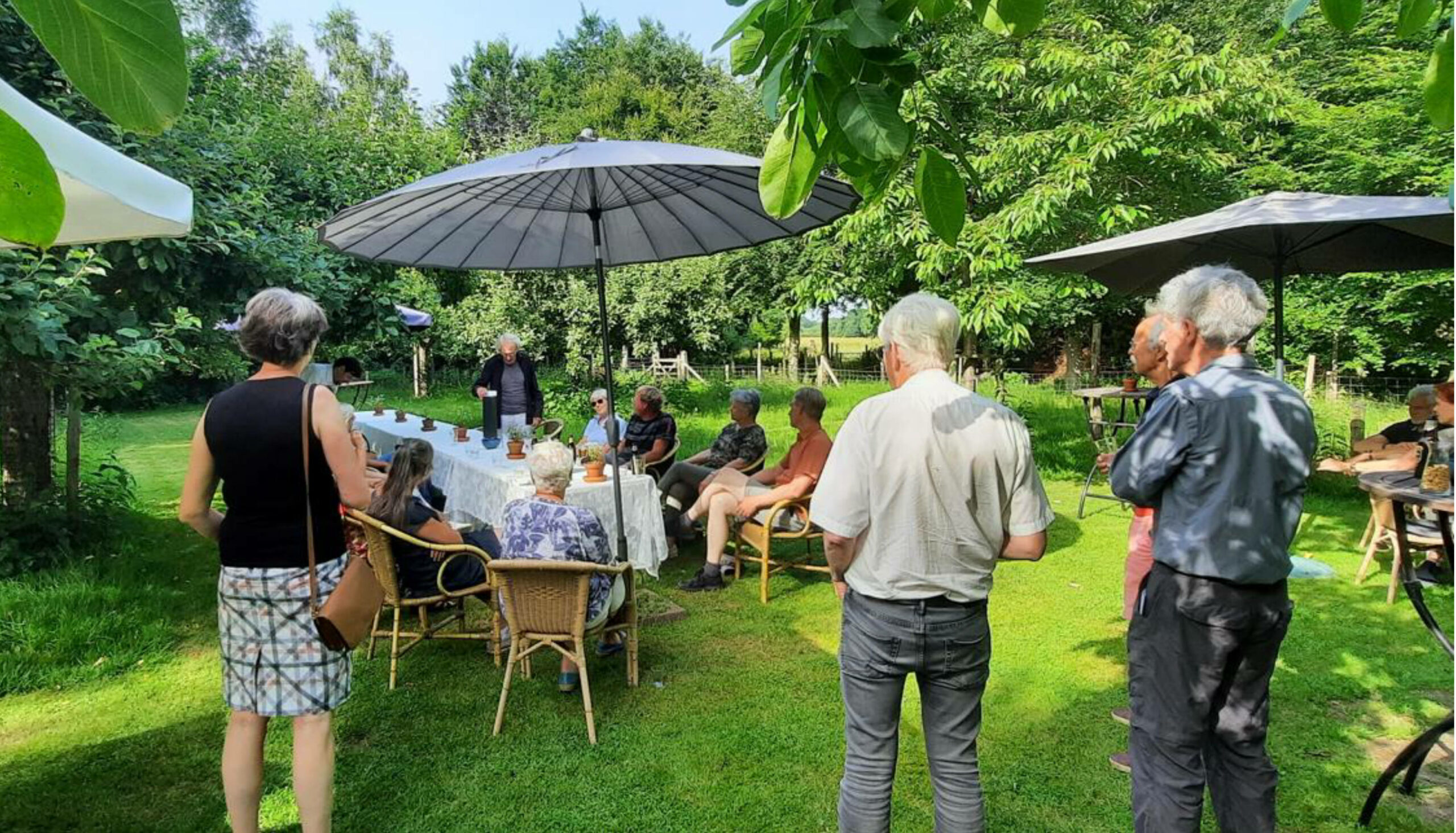 Groep mensen zit en staat rond een tafel onder parasols in een tuin.