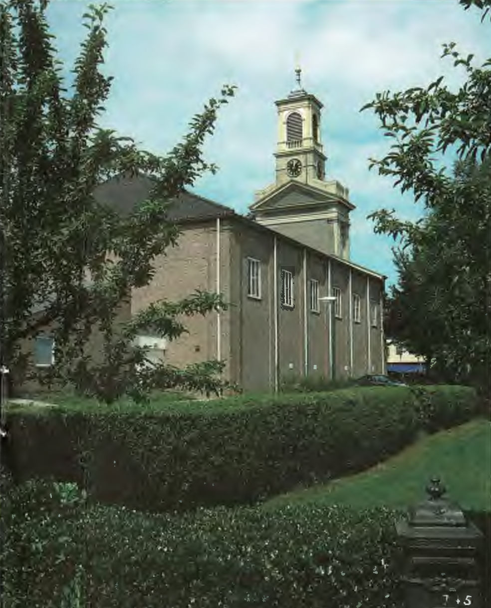 Een kerk met een kloktoren, omringd door bomen en heggen, tegen een bewolkte lucht.