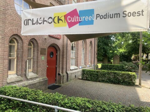 Bakstenen gebouw met rode deur en bordje "Artishock Cultureel Podium Soest", omgeven door groen.