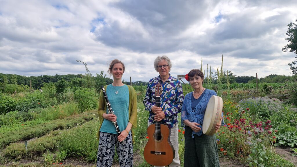 Drie mensen met muziekinstrumenten staan in een bloeiende tuin onder een bewolkte hemel.