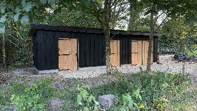 Zwarte houten schuur met bruine deuren, omgeven door bomen en planten.