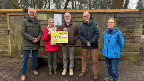 Vijf mensen staan voor een houten schuur. Twee van hen houden een grote cheque vast.