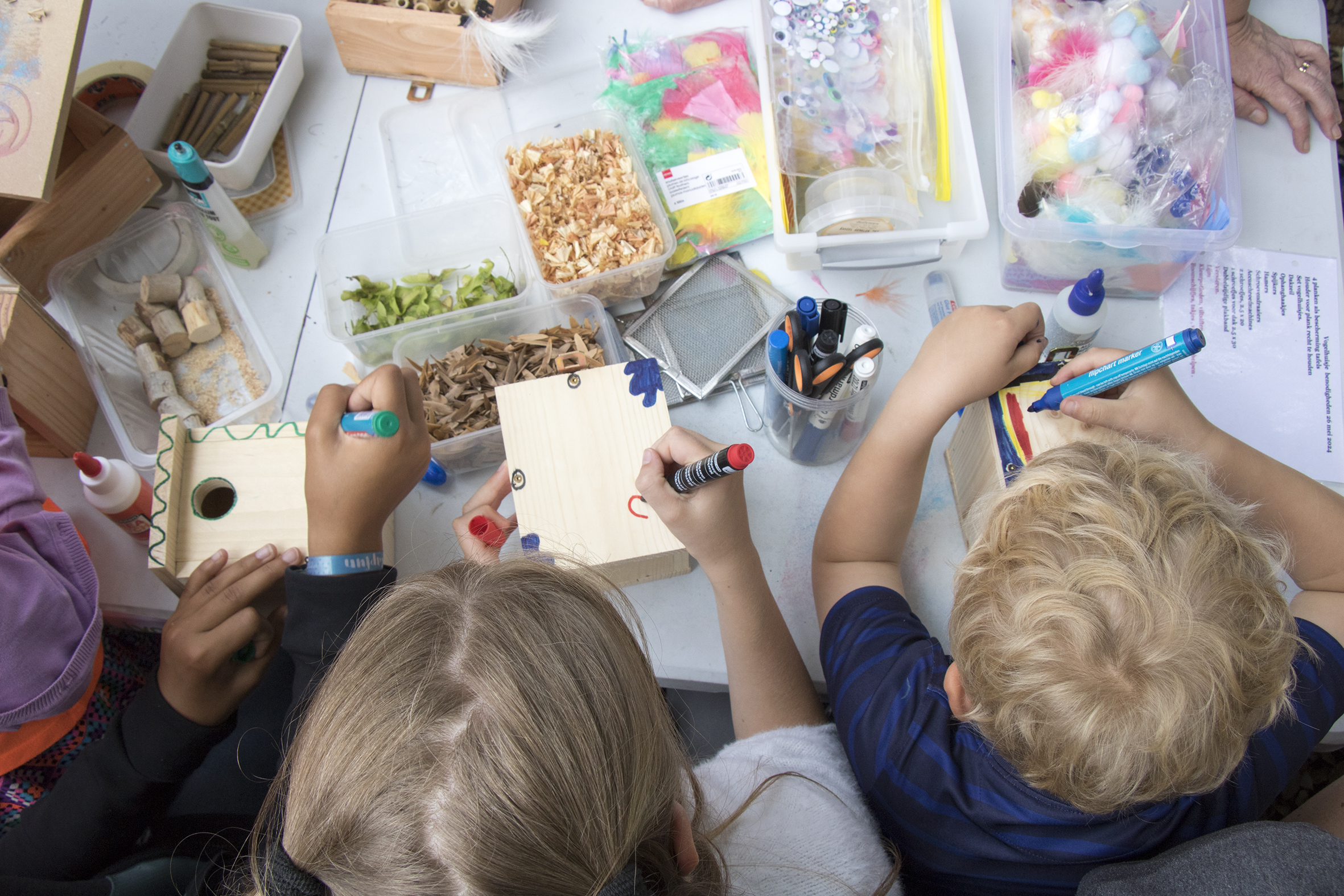 Drie kinderen versieren houten vogelhuisjes aan een tafel vol knutselmaterialen en stiften.